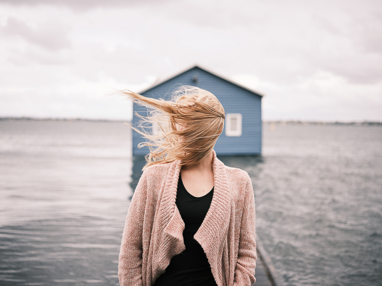 Woman with windblown hair standing by the water in front of a blue boathouse.