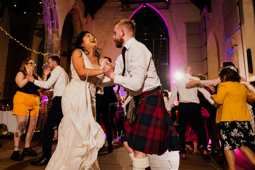 A joyful couple dancing at a wedding reception, with the groom wearing a traditional kilt.