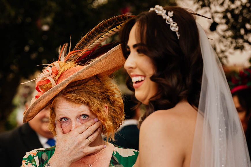 An emotional moment at a wedding, with a tearful woman in a hat embracing a smiling bride.