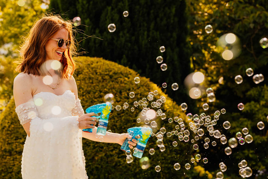 Woman in a white dress and sunglasses enjoying blowing bubbles in a sunny garden.