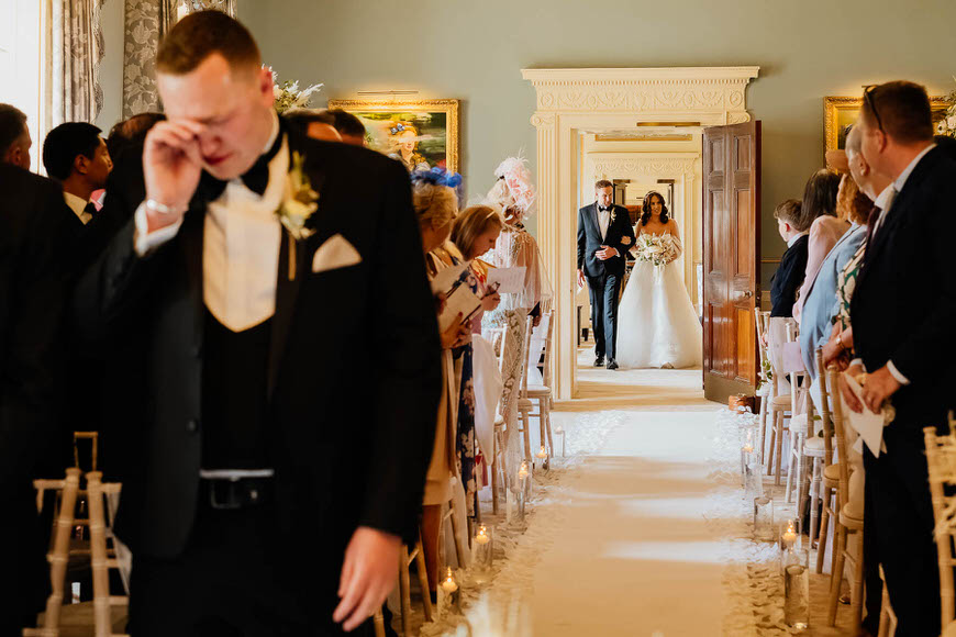 Emotional groom wiping away tears as the bride walks down the aisle during a wedding ceremony.