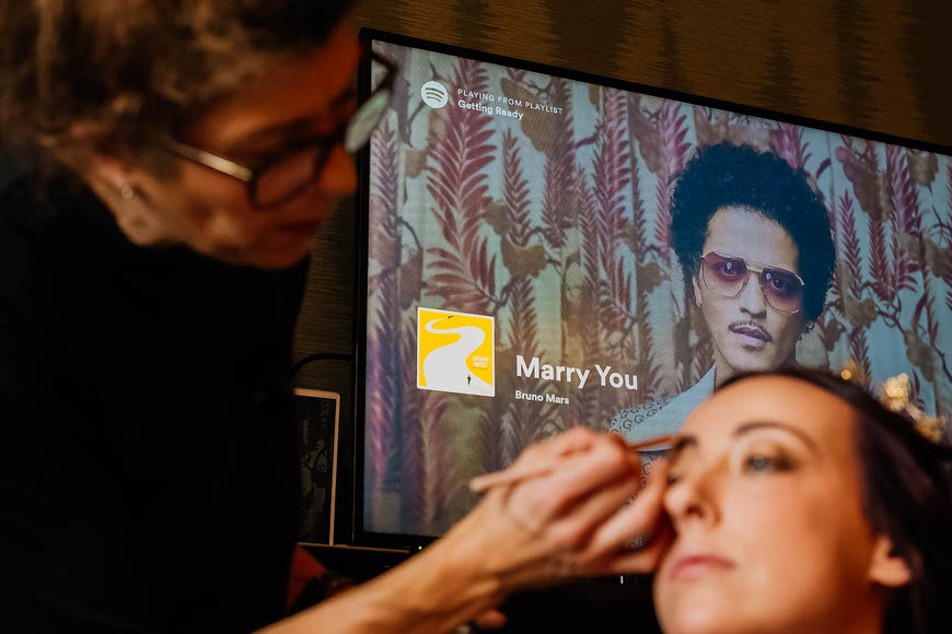 A makeup artist applies eyeshadow to a woman in front of a screen displaying a music track by bruno mars.