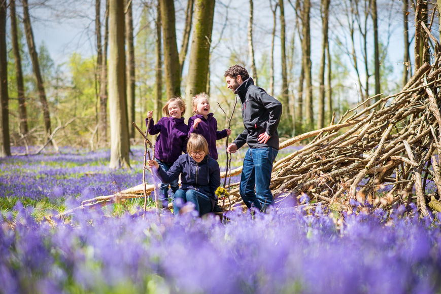 A family enjoys a day out in a blooming bluebell wood, with two children sitting on a wooden structure as the adults stand beside them smiling.