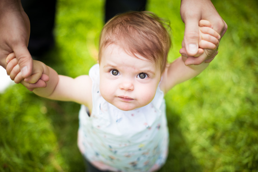 A baby with a focused expression being held up by the hands of an adult, standing against a background of green grass.