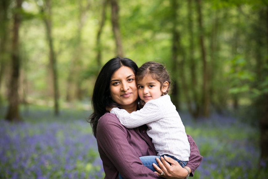 A woman holding a young child, both looking at the camera, with a backdrop of a forest floor covered in blue flowers.