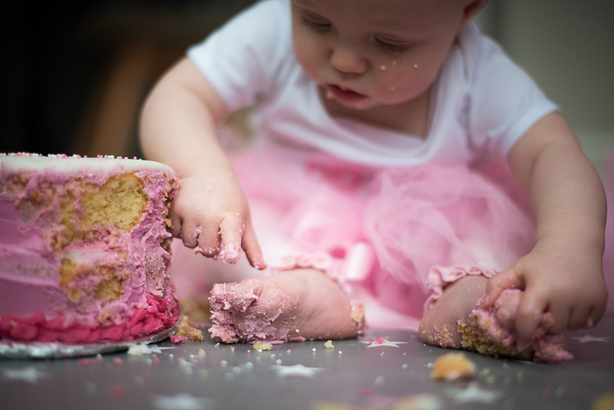A young child in a pink dress is exploring a pink birthday cake with her hands, making a bit of a mess with cake crumbs and frosting on her face and fingers.