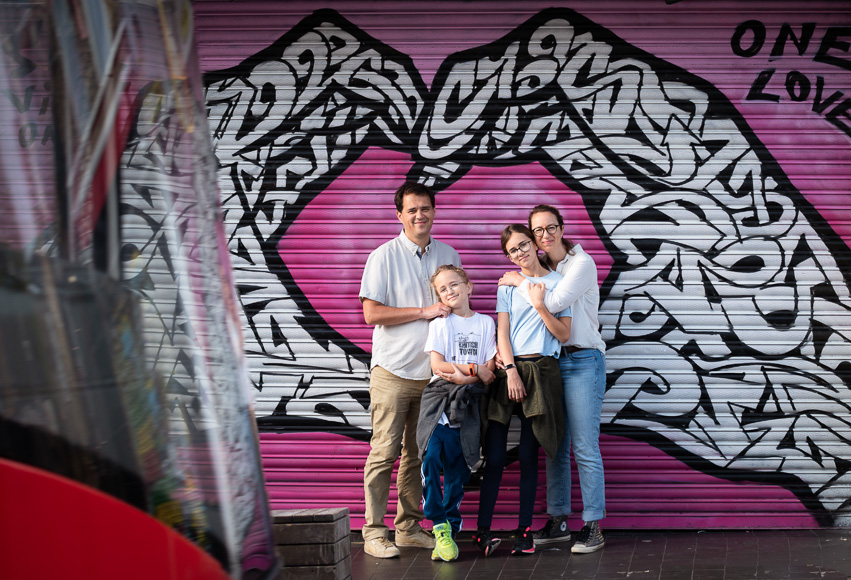 Three individuals, presumably a family, are standing in front of a vibrant graffiti wall with the words "one love" written on it. the youngest, a boy, is in the front and embraced by.