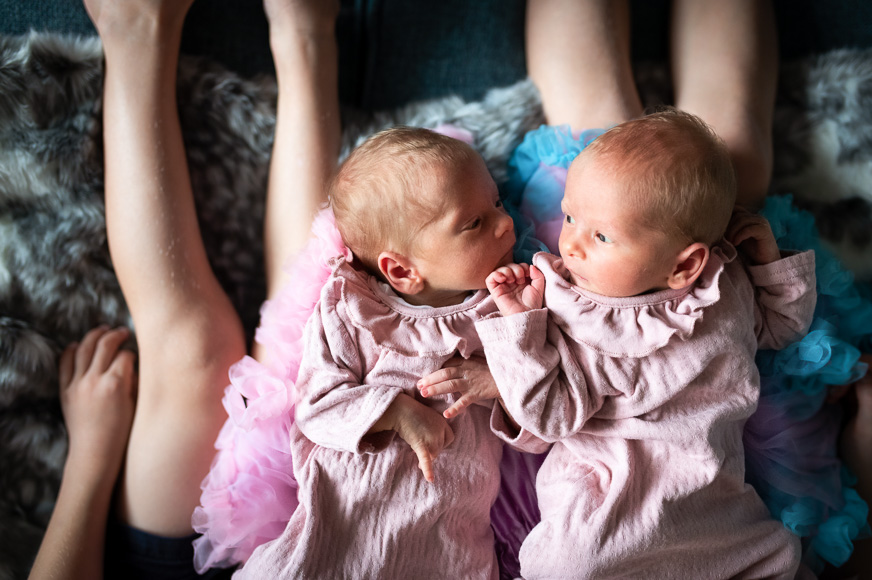 Two newborn babies dressed in ruffled pink outfits lie next to each other on a fuzzy surface, with a person's legs visible in the background.