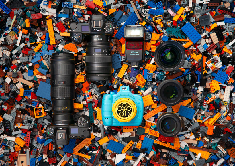 A collection of photography equipment, including cameras, lenses, and a flash, laid out on a colorful background of assorted lego bricks.
