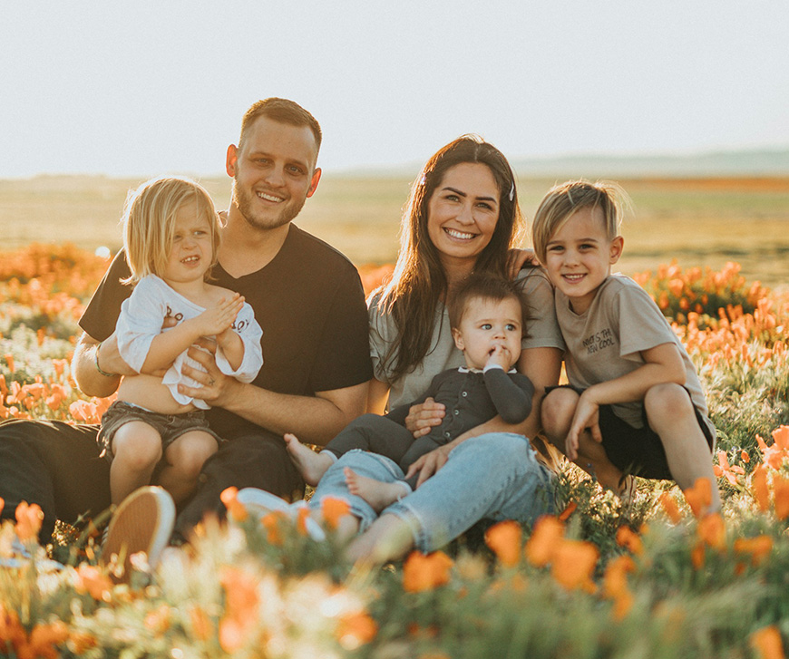 A family of five sits on a field with blooming orange flowers, smiling at the camera. The parents hold their three young children, two boys and one toddler, in their laps.