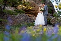 A couple in wedding attire sharing a tender moment amidst a natural landscape with rocks and wildflowers.