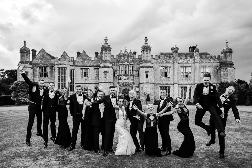 A joyful wedding party celebrating with a jump in front of an elegant, historic estate.