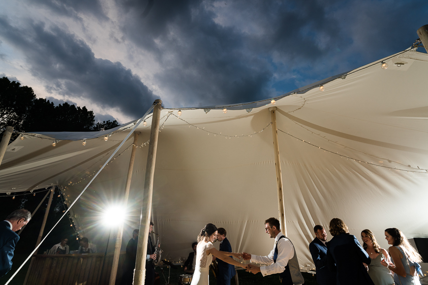 A gathering of people under a white tent adorned with string lights, with dramatic clouds in the sky above as the sun breaks through.