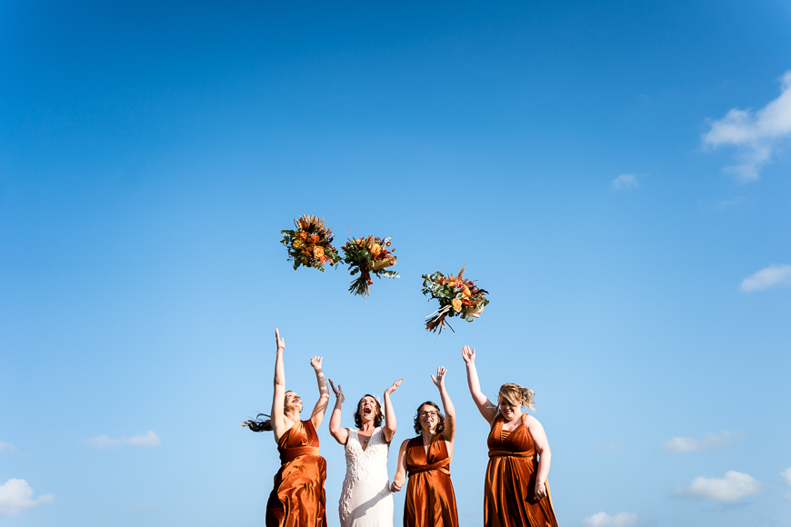 A bride and three bridesmaids, dressed in white and orange dresses respectively, joyfully toss bouquets into the air under a clear blue sky.