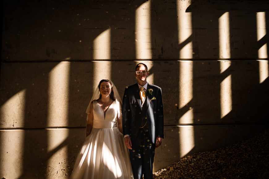 A bride and groom stand side by side, smiling, in a beam of sunlight creating strong contrasting shadows on a concrete wall behind them.