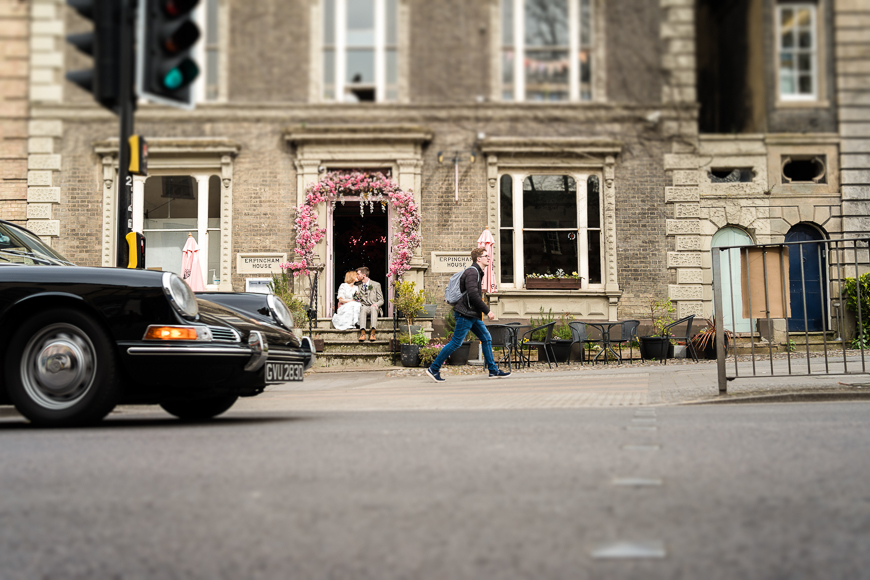 A person is walking across a street with a black car in the foreground and a quaint building with a flower-laden entrance in the background.