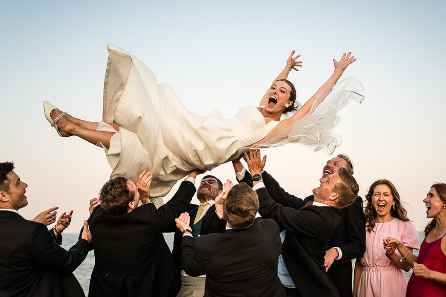 A bride in a white dress is joyfully being tossed into the air by a group of people, likely groomsmen and bridesmaids, during a wedding celebration.