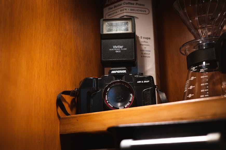 Vintage camera and external flash on a wooden shelf beside a coffee press and measuring guide.