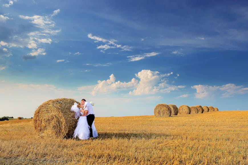 A couple in wedding attire embracing near a hay bale in a field.
