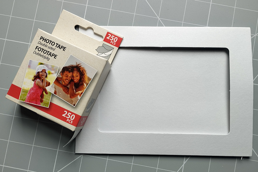 A box of photo tape next to an open empty scrapbook on a grey cutting mat.