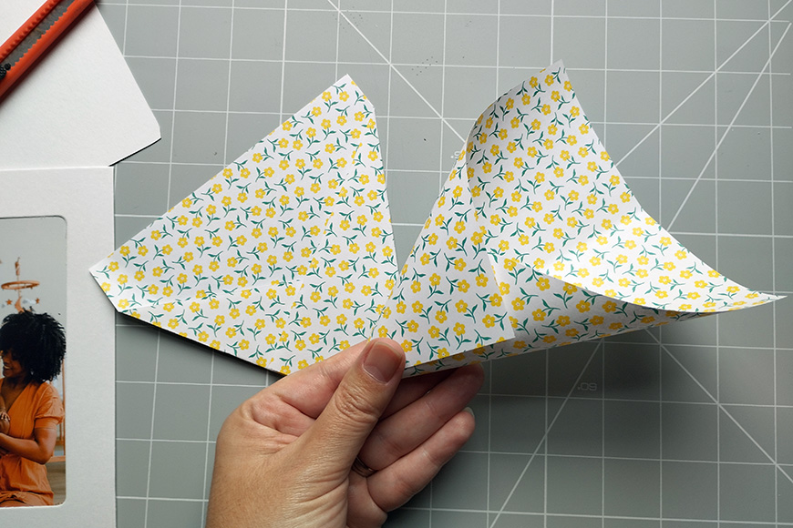 A hand holding a paper airplane with a floral pattern over a cutting mat.