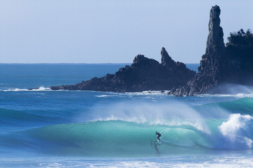 A surfer rides a large, curling wave with rocky outcrops in the background.