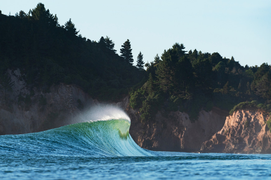 A wave breaking near a coastline with trees and cliffs in the background.