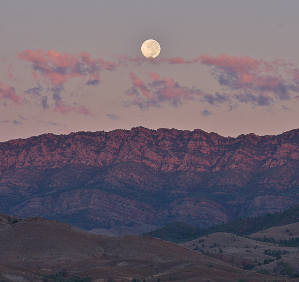Full moon rising above a rugged mountain range with pink-tinged clouds at dusk.