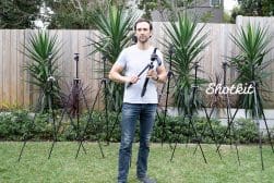 A man stands in a garden holding a camera tripod, surrounded by various tripods of different sizes, with plants in the background.
