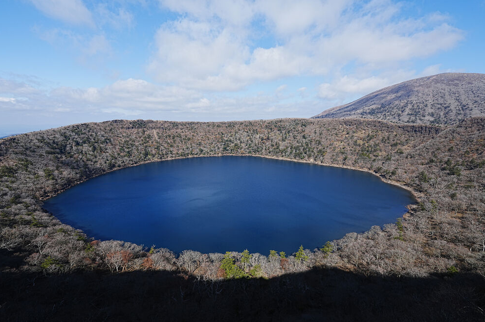 Aerial view of a circular blue lake surrounded by a forest in a volcanic crater, with a barren mountain slope in the background.