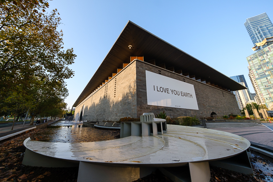 Modern building with a large banner stating "i love you earth" displayed on its facade, set against a clear sky with an urban park in the foreground.