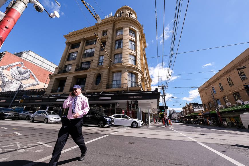 A person walking across a street corner with a historic building and tram lines overhead.