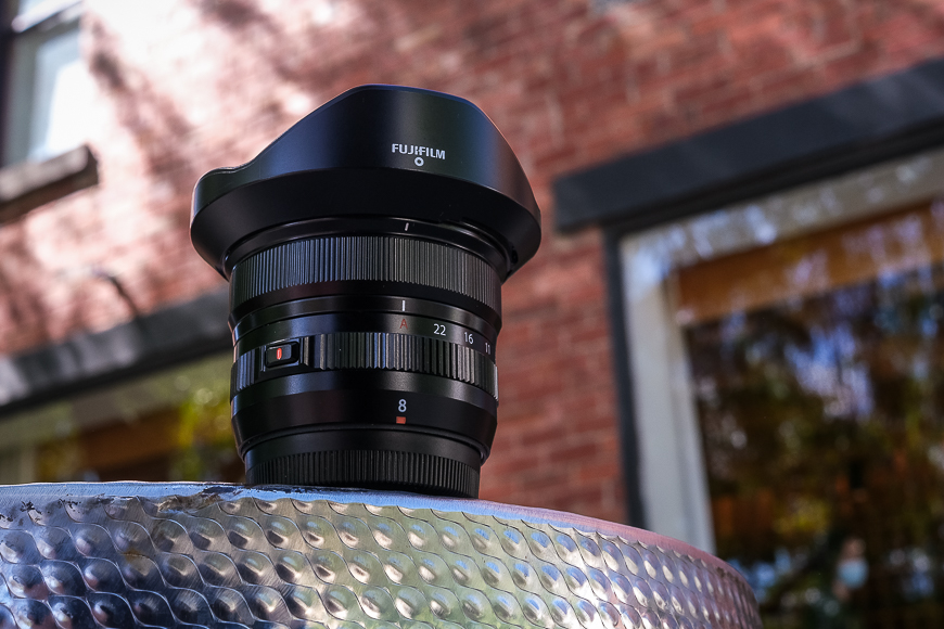 A fujifilm camera lens perched on a metal surface with a blurred brick background.