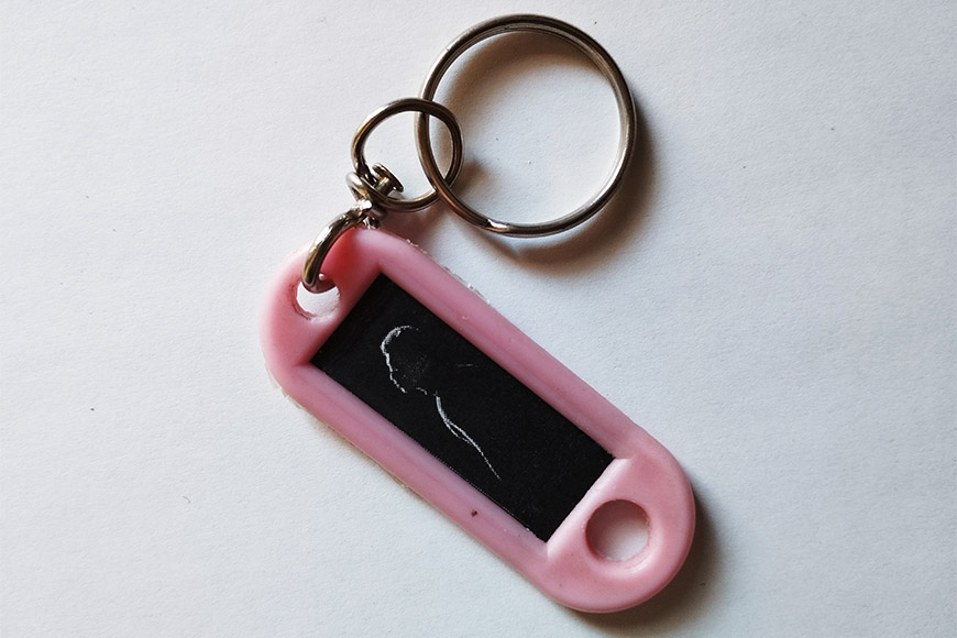 A pink keychain with a black chalkboard surface featuring a simple white chalk drawing of a cat.