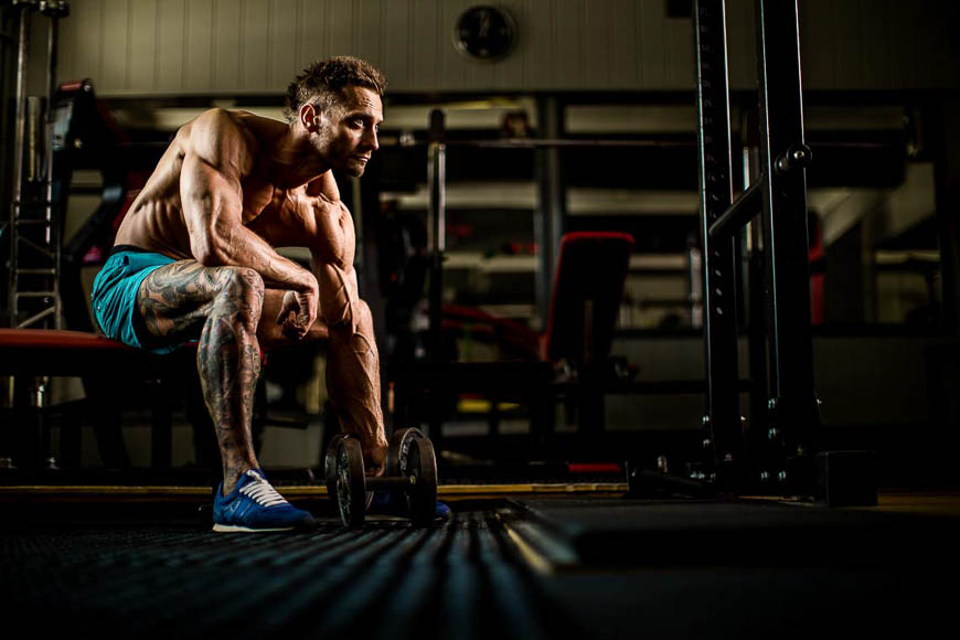 Muscular tattooed man sitting on a gym bench, resting and focusing on a dumbbell in a dimly lit gym.