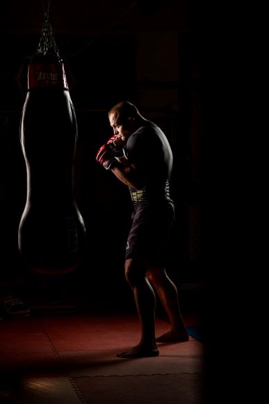 A man with boxing gloves stands focused beside a heavy bag in a dimly lit gym, highlighting his silhouette.