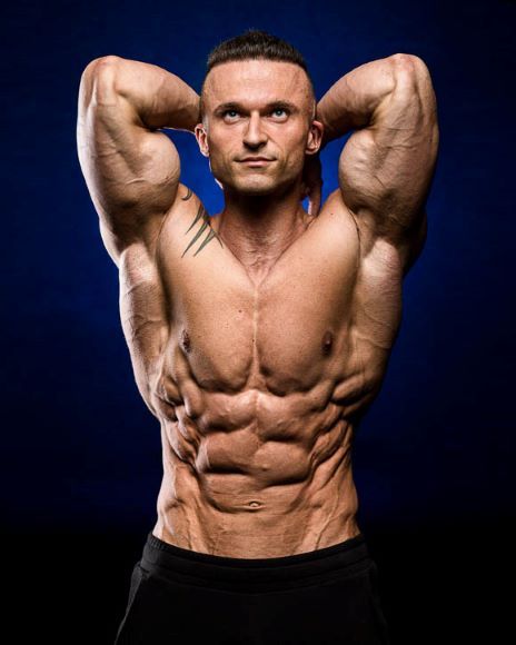 Muscular man posing with arms raised behind his head, showcasing his chiseled abs against a dark blue background.