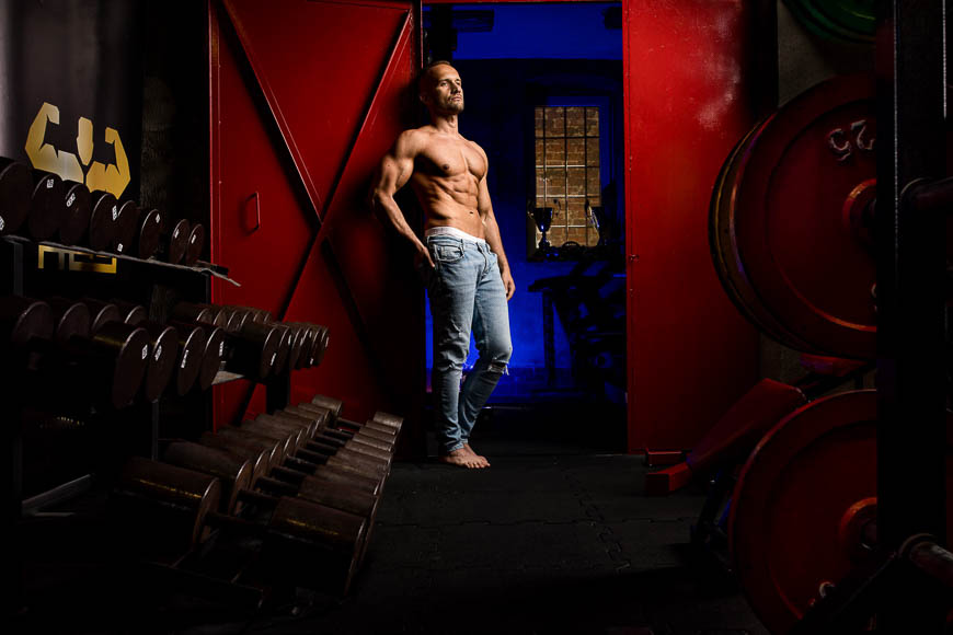 A shirtless muscular man in jeans standing confidently in a dimly lit gym with weight racks and red walls.