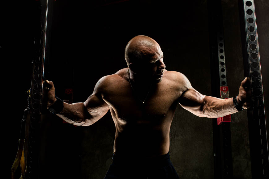 Muscular bald man flexing arms dramatically in a dark gym, illuminated by a stark overhead light.