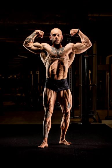 A muscular bald man with tattoos flexing his muscles in a gym, wearing black shorts.