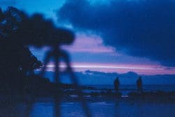 Blurred silhouette of a camera on a tripod with two people standing by a beach at dusk under a colorful sky.