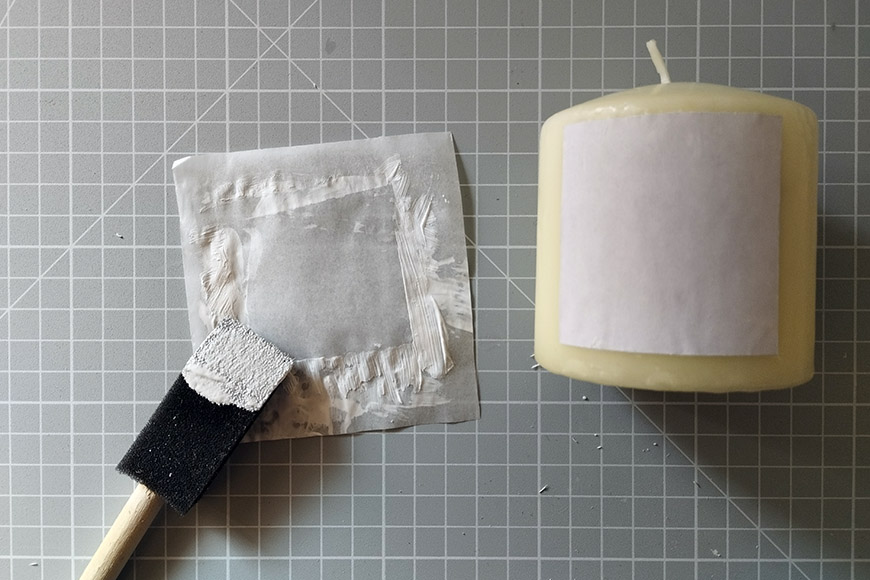 A paintbrush with a sponge applicator next to a white candle with a safety label, both placed on a gray grid cutting mat.