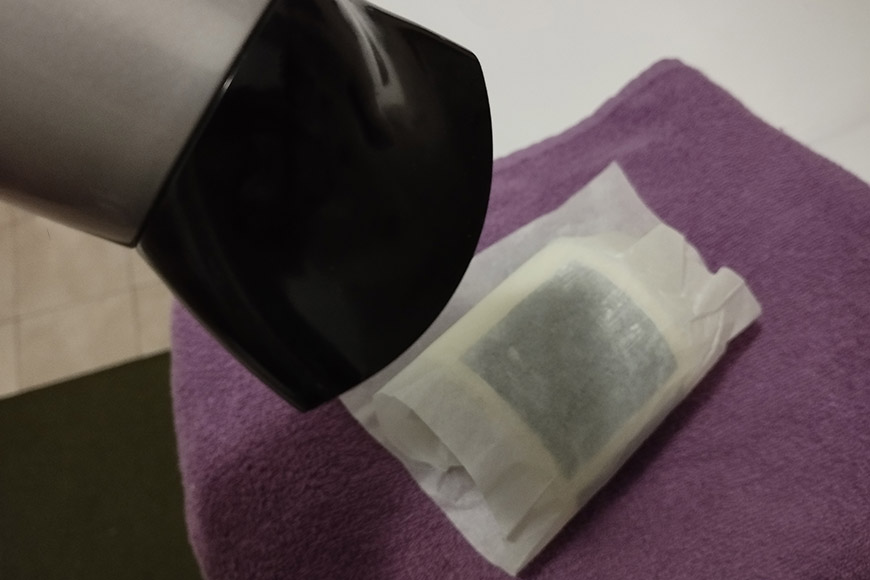 A hairdryer directed towards a tea bag resting on a purple towel, indicating an attempt to dry or heat the tea bag.