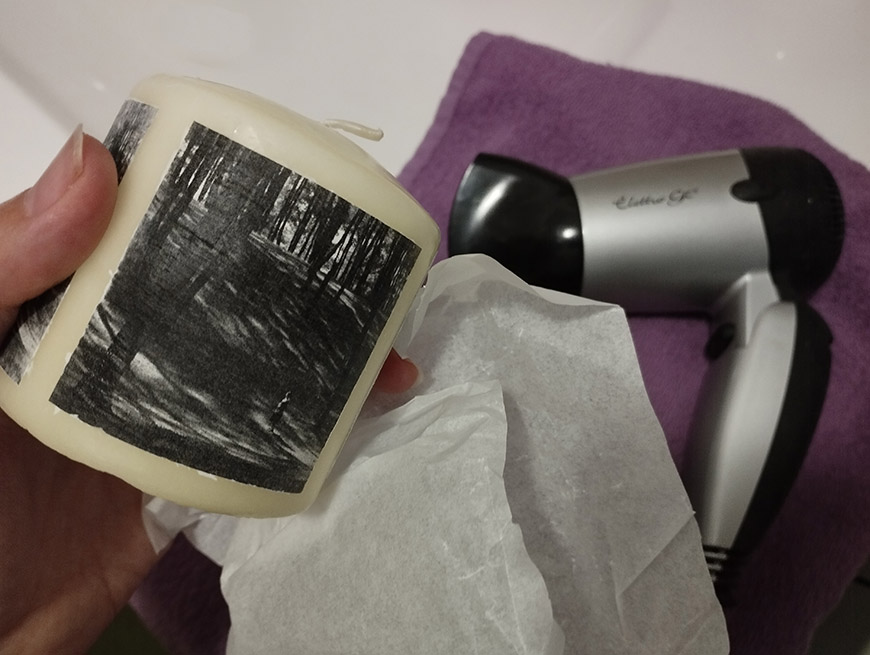A hand holding a homemade candle with a black and white photo transfer on its surface, next to a hairdryer and a purple towel.