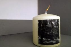 A lit cylindrical candle with a monochrome forest scene printed on its side, placed on a gray surface against a two-tone gray background.