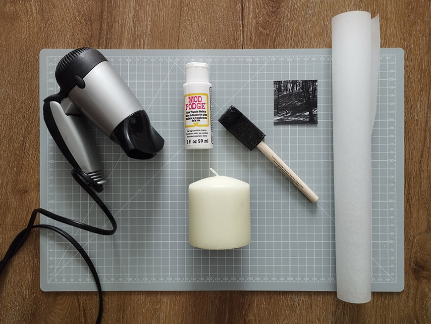 Craft supplies including a heat gun, mod podge glue, paintbrush, candle, and parchment paper on a wooden surface.