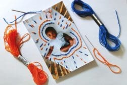 A photo of a baby surrounded by colorful thread embroidery in progress on paper, with needles and threads in orange and blue on the side.