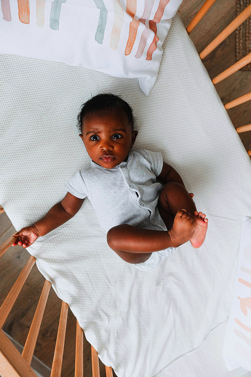A baby with dark eyes and short hair lies on a white patterned blanket in a wooden crib, looking up.