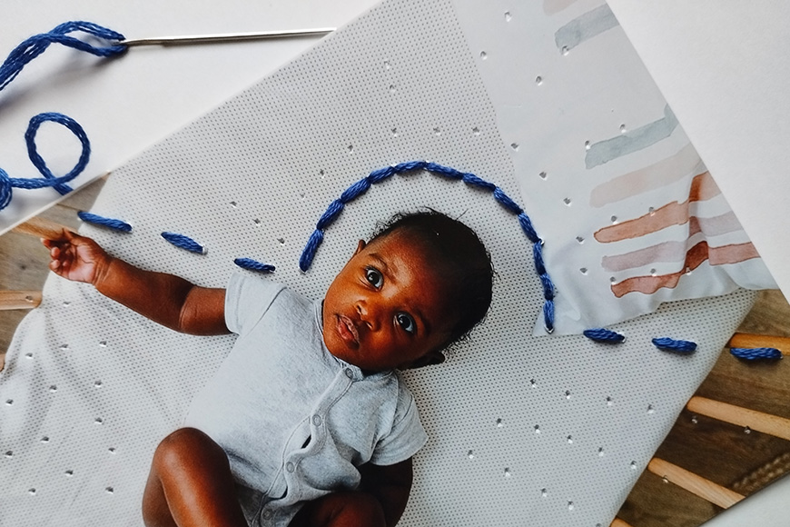 A baby lying on a mat next to a semi-completed embroidery project with blue yarn.