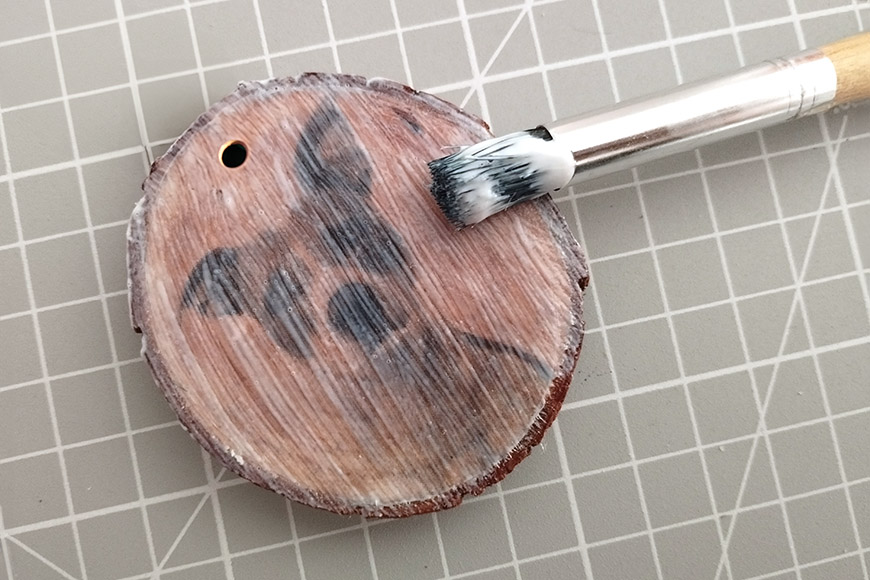 A paintbrush with white bristles applying gray paint to a round wooden disc on a grid-patterned surface.
